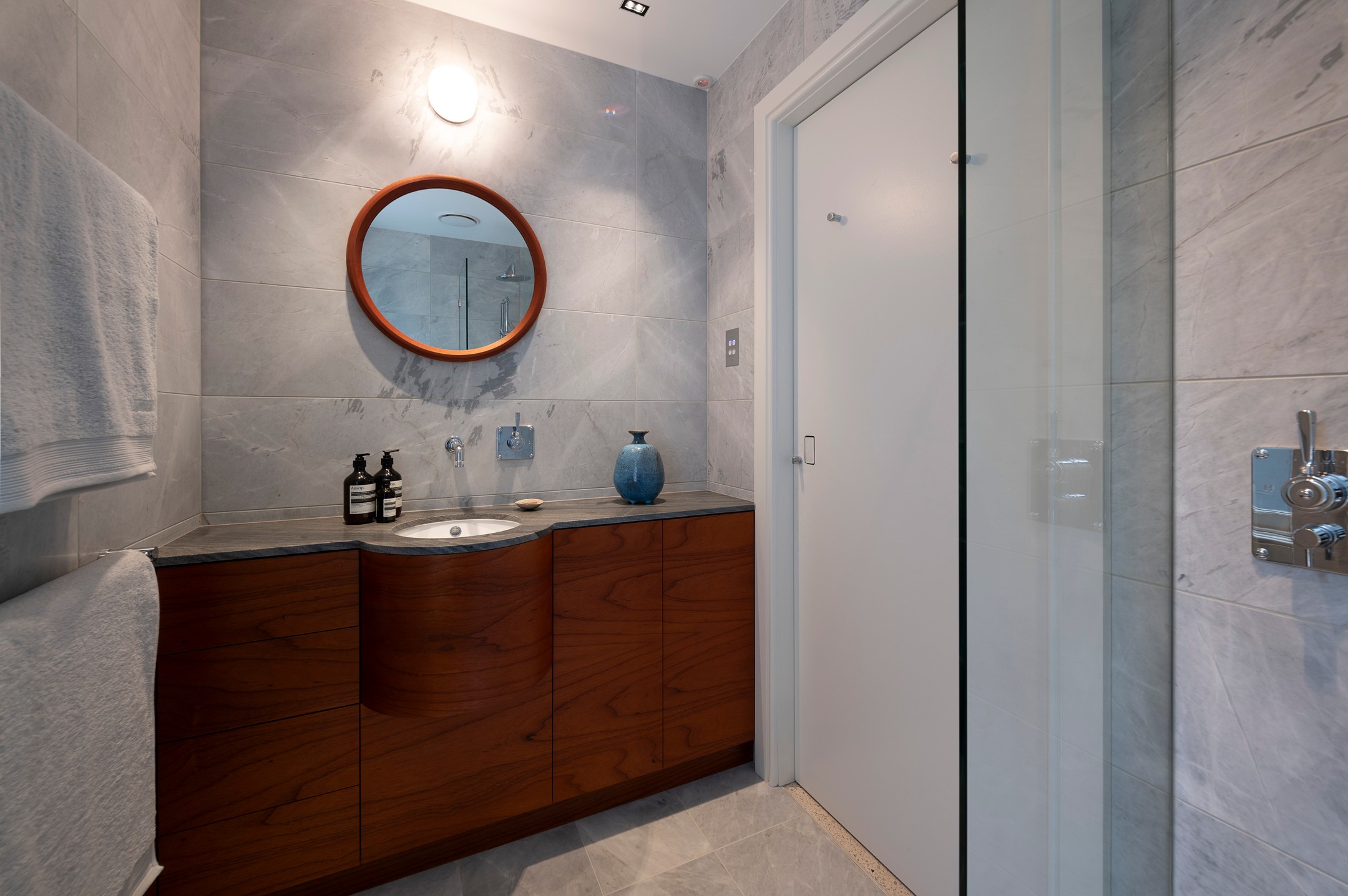 Bathroom with a curved timber vanity, stone benchtop, round timber-framed mirror and marble tiles