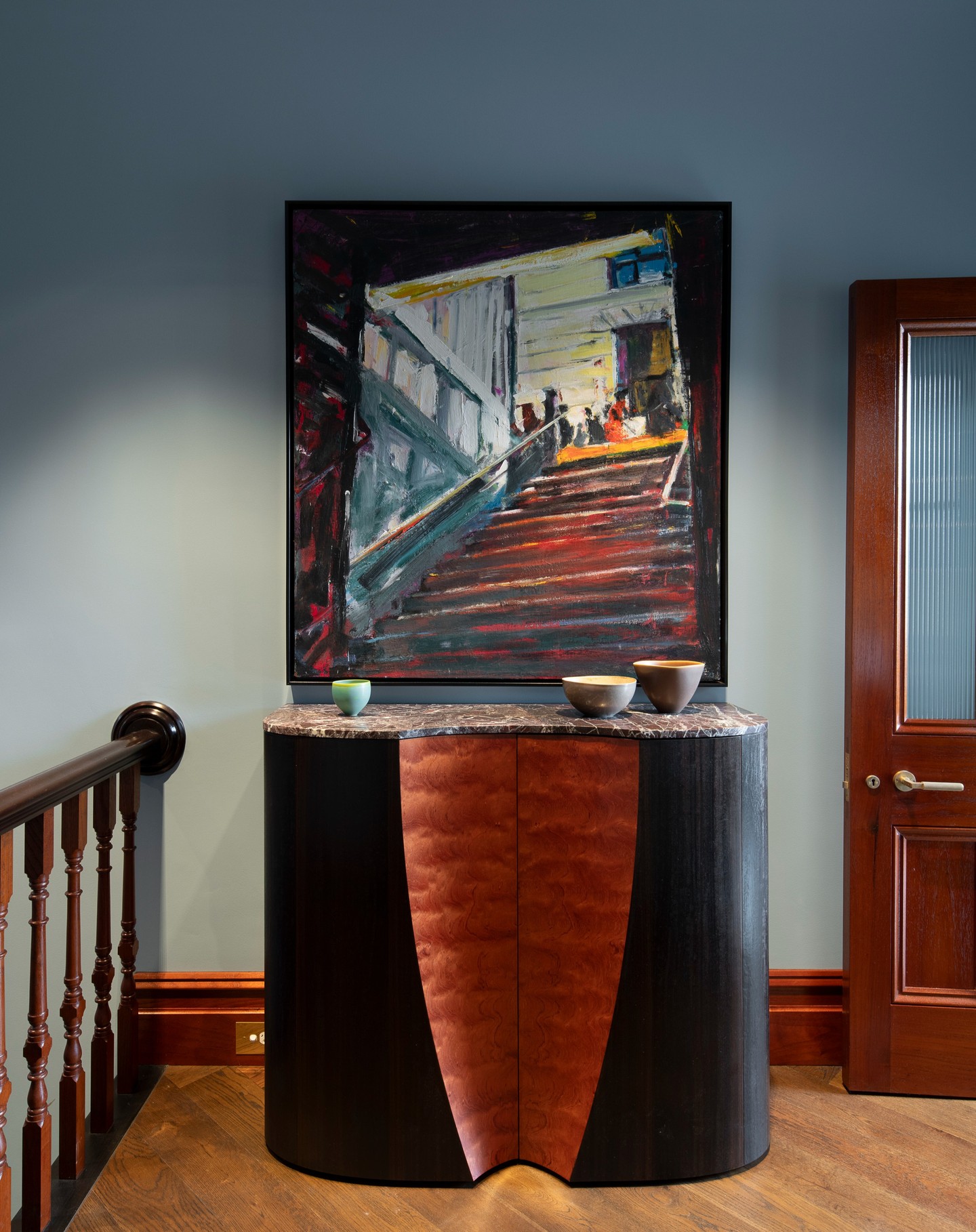 Curved dark timber cabinet with a book-matched veneer front and marble top beneath a framed painting