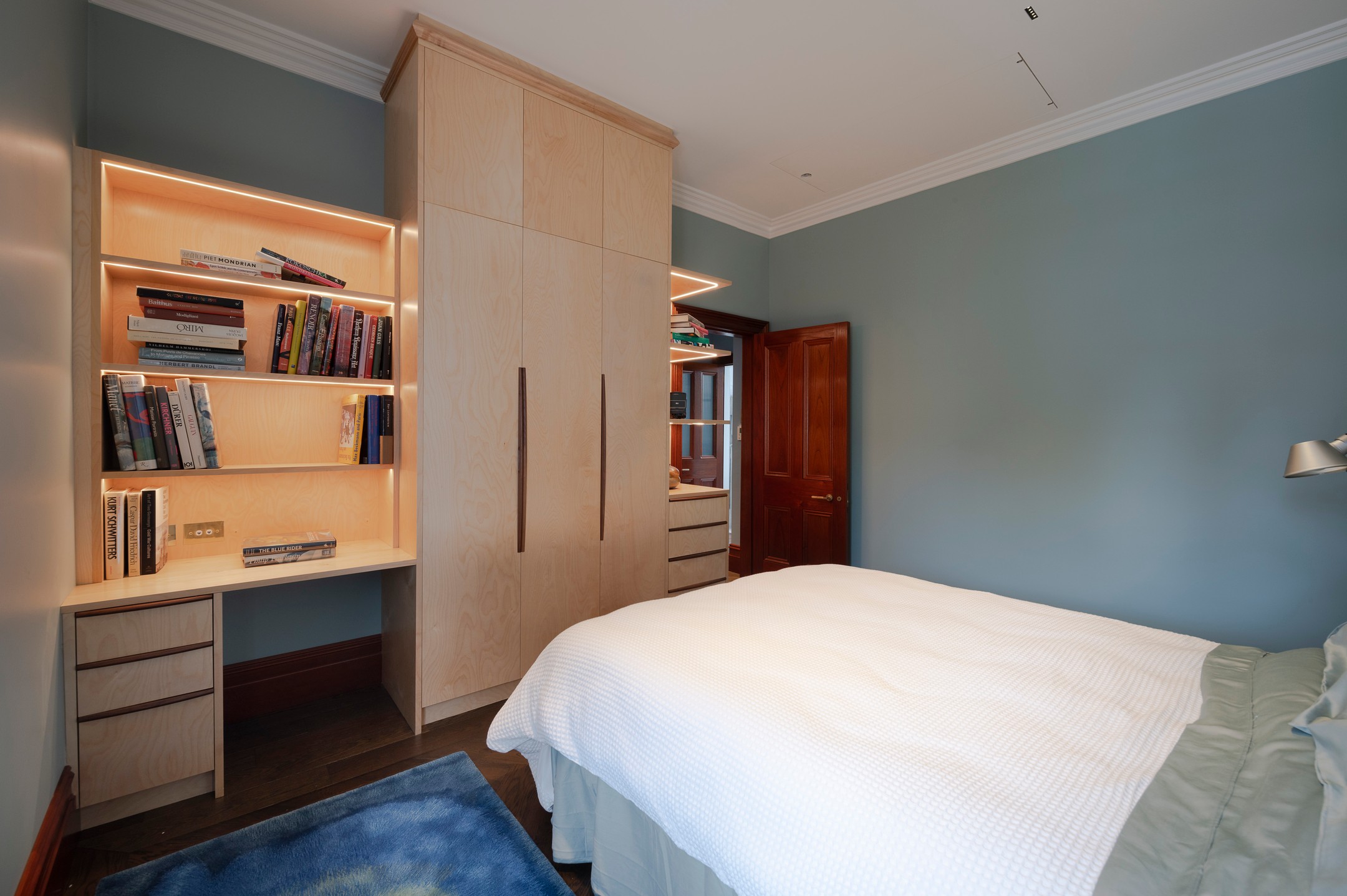 Birch ply wardrobe and illuminated built-in bookshelves with desk in a bedroom